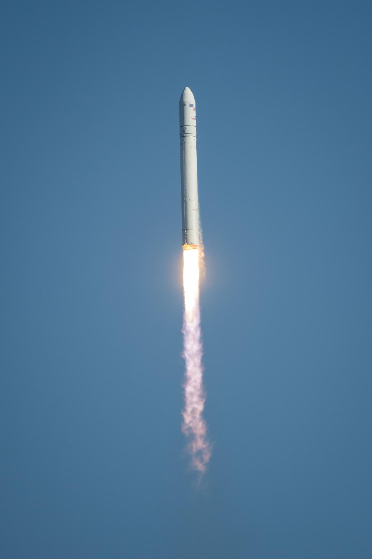 青空を背景に、白いロケットが点火した主エンジンから炎と煙を噴き出しながら打ち上げられている様子。